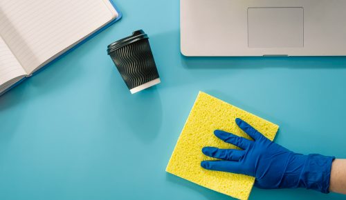 A hand in a rubber glove wipes a workplace with a laptop with a rag, flat lay.